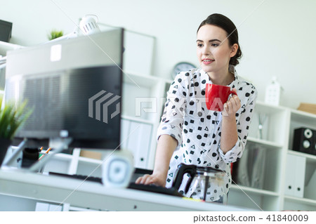 A young girl is standing in the office near the table, holding a red mug in her hand and typing on 41840009