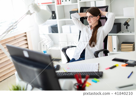 A young girl is sitting at a table in the office, laying her hands behind her head and looking at 41840210