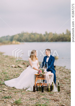 Happy newlyweds are holding hands while sitting at the wedding table on the coast. Vertical photo. 41867271