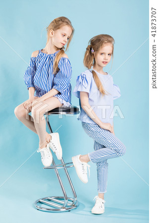 Girls twins in light blue clothes are posing near a bar stool on a blue background. Girls twins in light blue clothes are posing near a bar stool on a blue background. 41873707