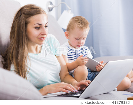 Smiling mom is productively working behind laptop while child playing on tablet Smiling mom is productively working behind laptop while child playing on tablet 41879383