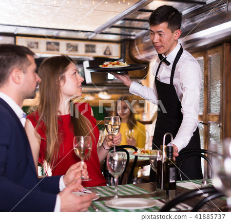 Man waiter is brings order to couple who have dinner and drink wine Man waiter is brings order to couple who have dinner and drink wine 41885737