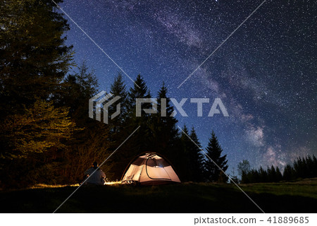 Male hiker enjoyng night camping near tourist tent at campfire under blue starry sky and Milky way 41889685