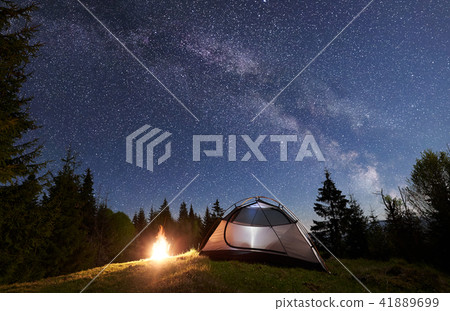 Night camping in mountains. Tourist tent by campfire near forest under blue starry sky, Milky way 41889699
