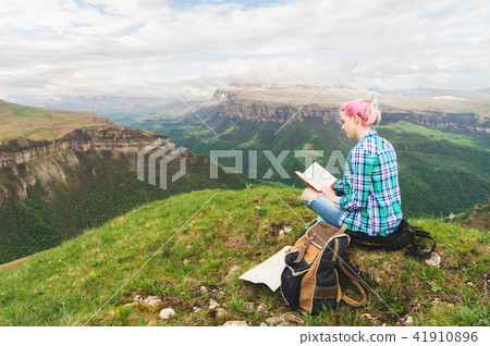 A traveler girl sits in the mountains on the grass and reads a book on the background of epic A traveler girl sits in the mountains on the grass and reads a book on the background of epic 41910896