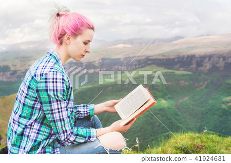A traveler girl sits in the mountains on the grass and reads a book on the background of epic A traveler girl sits in the mountains on the grass and reads a book on the background of epic 41924681