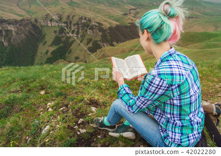 A traveler girl sits in the mountains on the grass and reads a book on the background of epic A traveler girl sits in the mountains on the grass and reads a book on the background of epic 42022280