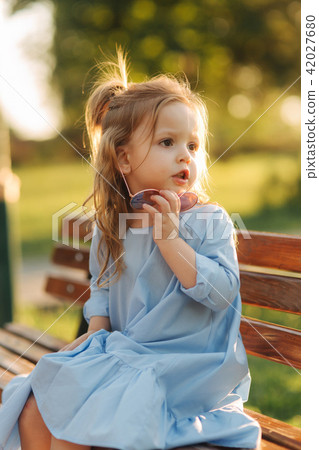 Little girl model in blue dress and sun glasses sits on a bench in the park 42027680
