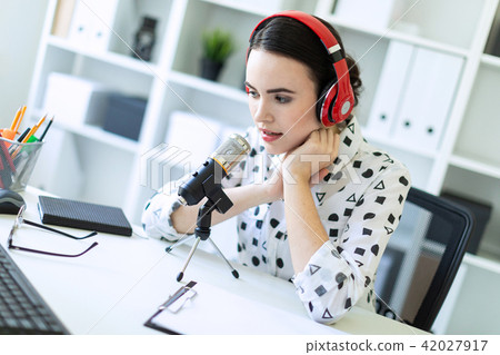 Beautiful young girl sitting in headphones and with microphone at table in office and talking. 42027917