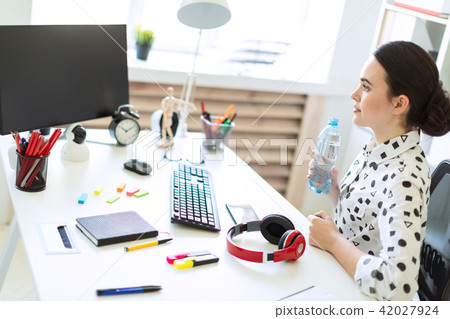 A beautiful young girl sits at a table in the office and holds a bottle of water in her hands. 42027924