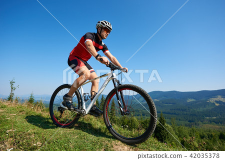 Young male sportsman cyclist in helmet, sunglasses and full equipment riding bike on grassy hill Young male sportsman cyclist in helmet, sunglasses and full equipment riding bike on grassy hill 42035378
