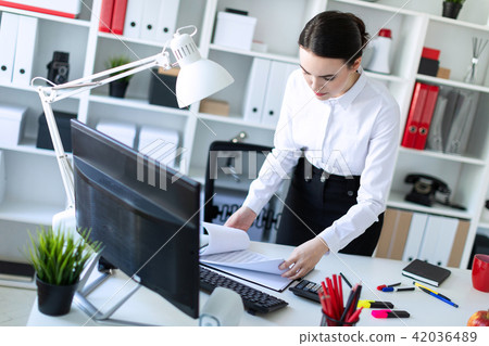 A young girl in the office is standing near the table and looks through the documents. 42036489