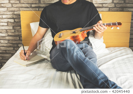 Happy young asian man playing ukulele sitting on bed in bedroom. 42045440