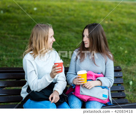 Two girl friends. In the summer on bench. They talk with hands holding cups of coffee and tea. A 42083418