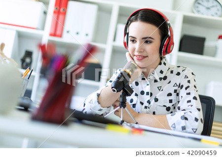 Beautiful young girl sitting in headphones and with microphone at table in office and talking. 42090459