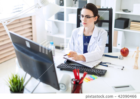 A beautiful young girl in a white robe is sitting at a computer desk with documents and a pen in her 42090466
