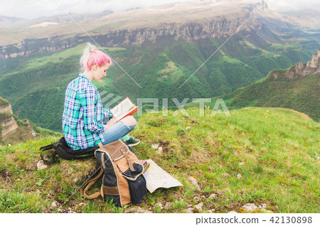 A traveler girl sits in the mountains on the grass and reads a book on the background of epic 42130898