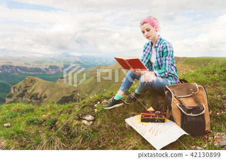 A traveler girl sits in the mountains on the grass and reads a book on the background of epic A traveler girl sits in the mountains on the grass and reads a book on the background of epic 42130899