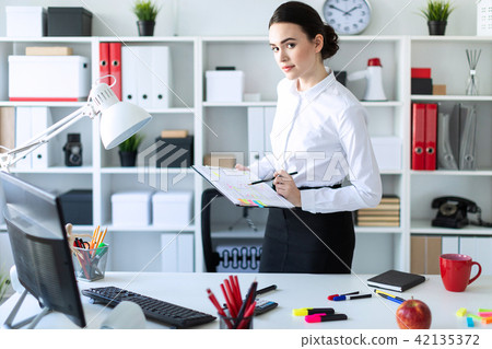 A young girl in the office is standing near the table and holds in her hands a sheet for notes and a 42135372