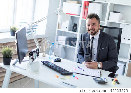 A man is sitting in the office at the table and holding a glass of coffee. 42135373