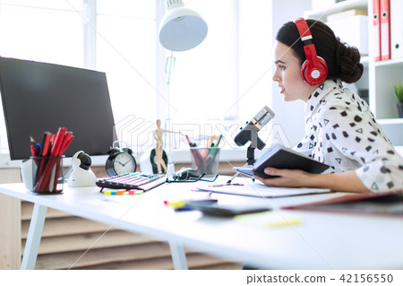 Beautiful young girl sits in headphones and with a notepad at the table in the office and speaks 42156550