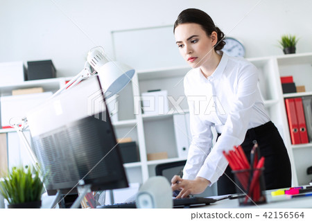 A young girl in the office is standing near the table, holding a pencil in her hand and typing text 42156564