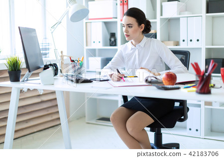 A young girl in the office holds a pink marker in her hand and works with a computer and documents. 42183706