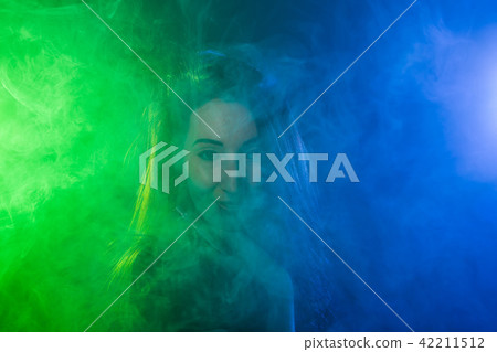 Portrait of young woman in neon blue and green smoke with vape or e-cigarettes Portrait of young woman in neon blue and green smoke with vape or e-cigarettes 42211512