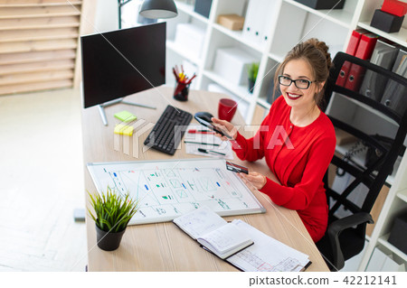 A young girl is sitting at the desk in the office, holding a bank card and phone in her hand. A young girl is sitting at the desk in the office, holding a bank card and phone in her hand. 42212141