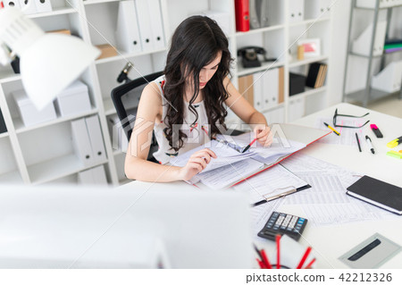 A beautiful young girl sits at an office desk is holding a pen in her hand and thumbs through a 42212326