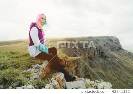 A girl-traveler with multicolored hair sits on the edge of a cliff and looks to the horizon on a 42241323