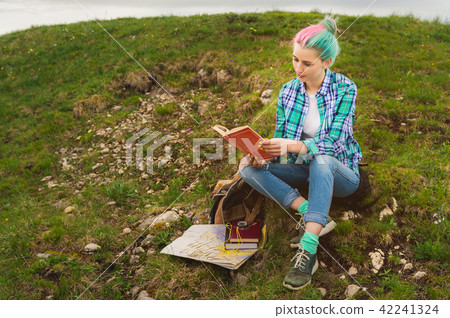A traveler girl sits in the mountains on the grass and reads a book on the background of epic 42241324