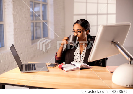 Afroamerican businesswoman sitting by the table in her officce and drinling coffee or tea. Afroamerican businesswoman sitting by the table in her officce and drinling coffee or tea. 42286791
