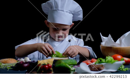 Little boy is cooking a burger at the kitchen, he is wearing chefs suit 42299213