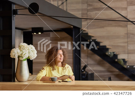 Siling curly girl looking at side, keeping blocknote sitting on wooden table. Siling curly girl looking at side, keeping blocknote sitting on wooden table. 42347726