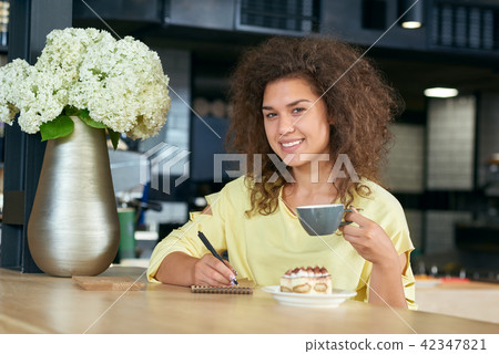 Smiling girl with curly hair drinking coffee, writing notes. Smiling girl with curly hair drinking coffee, writing notes. 42347821