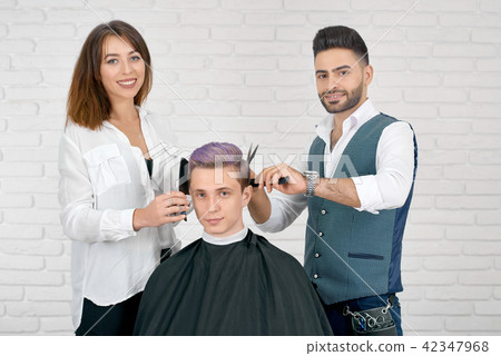 Female and male hairstylists doing haircut for young client with toned hair. 42347968