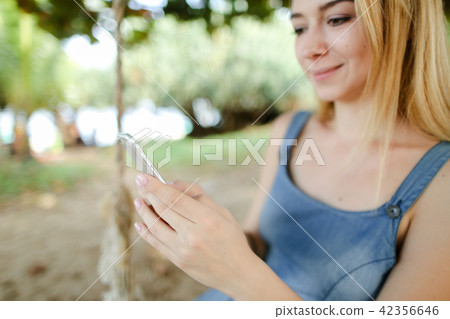 Young nice girl riding swing and using smatrphone, sand and tree in background. Young nice girl riding swing and using smatrphone, sand and tree in background. 42356646