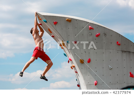 Photo of young sporty brunet in red shorts hanging on wall for rock climbing against blue sky with Photo of young sporty brunet in red shorts hanging on wall for rock climbing against blue sky with 42377974
