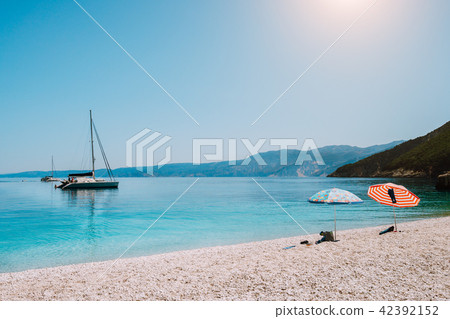 Idyllic white beach with umbrellas on lazy summer day. Sailing boat on calm crystal clear water Idyllic white beach with umbrellas on lazy summer day. Sailing boat on calm crystal clear water 42392152