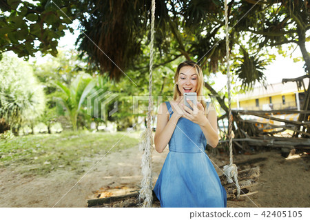 Young blonde girl chatting by smartphone and riding swing in Thailand, sand in background. Young blonde girl chatting by smartphone and riding swing in Thailand, sand in background. 42405105
