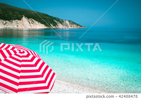 Striped red sun beach umbrella on a pebble beach with turquoise blue sea, white rocks and sky in 42408478