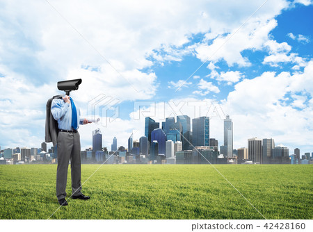 Camera headed man standing on green grass against modern cityscape 42428160