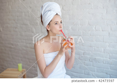 Caucasian smiling woman after spa covered with white towel drinking refreshing drink in glass bottle 42473597