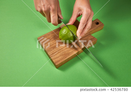 A woman's hands cut a natural green ripe lime in half on a wooden cutting board on a green A woman's hands cut a natural green ripe lime in half on a wooden cutting board on a green 42536780