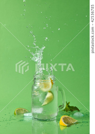 Cocktail splash from ice cube in the glass jar. Drops of liquid and pieces of lemon and lime on a 42536785