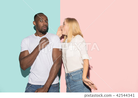 The young couple whispering a secret behind her hand over studio background The young couple whispering a secret behind her hand over studio background 42544341