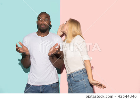 The young couple whispering a secret behind her hand over studio background The young couple whispering a secret behind her hand over studio background 42544346
