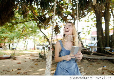 Young smiling girl using tablet and riding swing on sand, wearing jeans sundress. 42571286