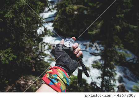 A gloved hand holds a magnetic compass against the background of a coniferous forest and mountain A gloved hand holds a magnetic compass against the background of a coniferous forest and mountain 42578003